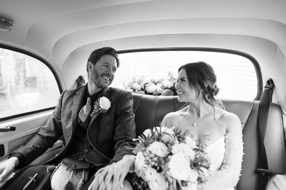 A bride and groom sitting in the back of a car, smiling at each other, with the bride holding a bouquet of flowers at their Edinburgh elopement