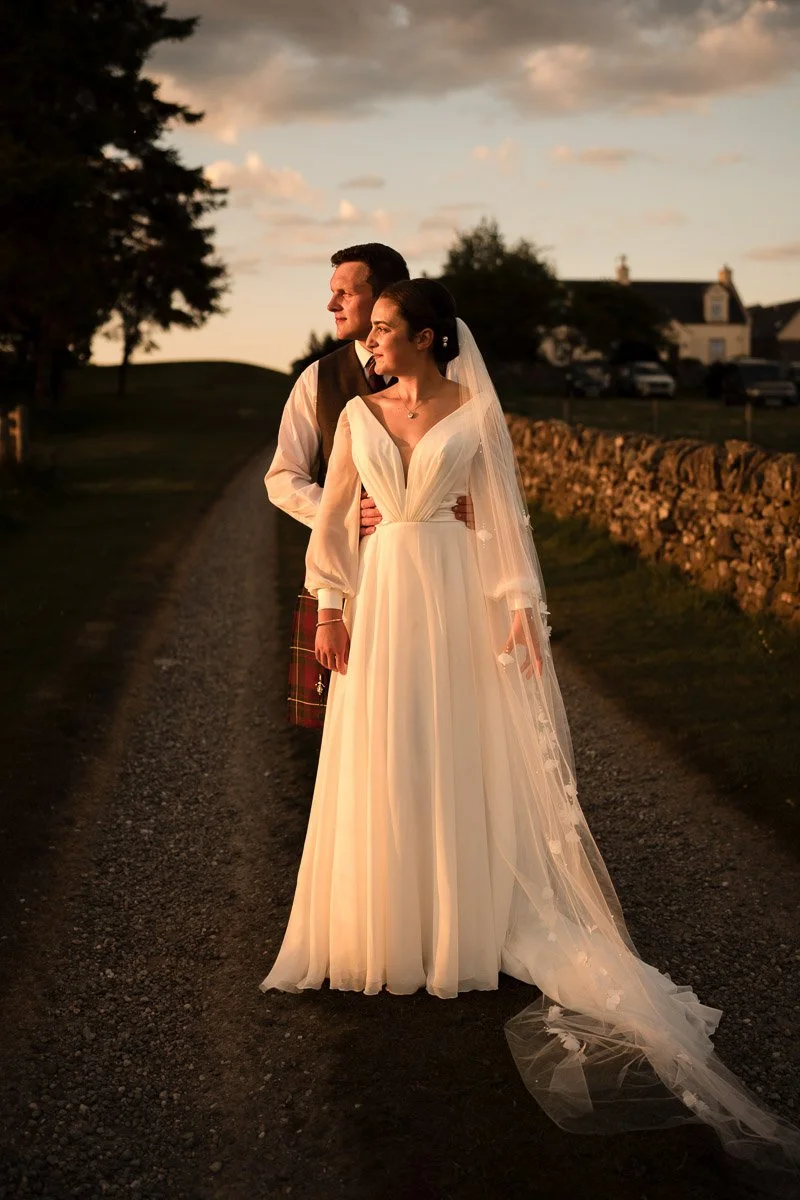A bride and groom standing together on a rural dirt road during sunset at their wedding at Ardoch Loch Lomond. The bride is wearing a flowing white wedding gown with a veil, and the groom is dressed in traditional Scottish attire.