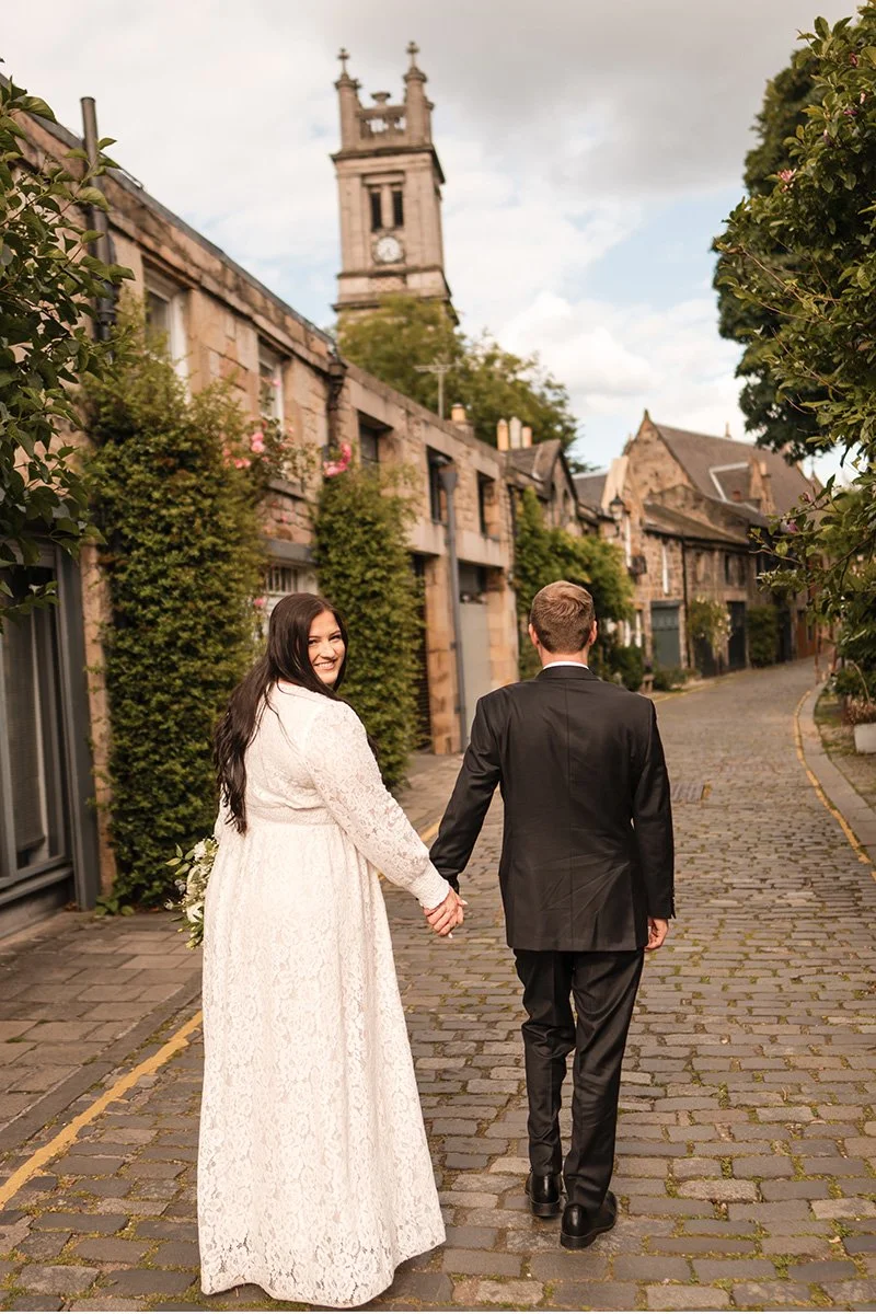 A bride and groom holding hands and walking down a cobblestone street in a small town, with a clock tower in the background on Circus Lane after their Edinburgh city elopement