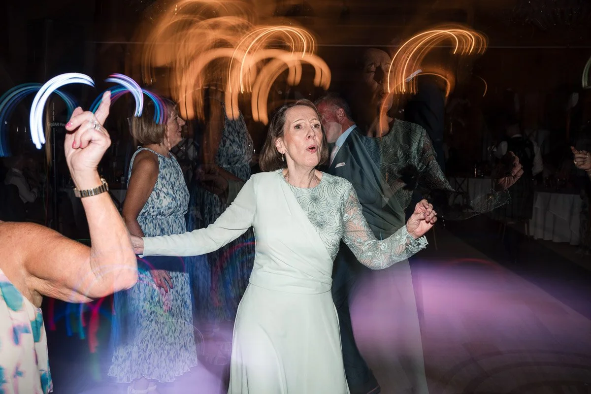 An older woman in a dress with lace sleeves is dancing at a wedding, with colourful light streaks and other people dancing in the background at Boturich Castle