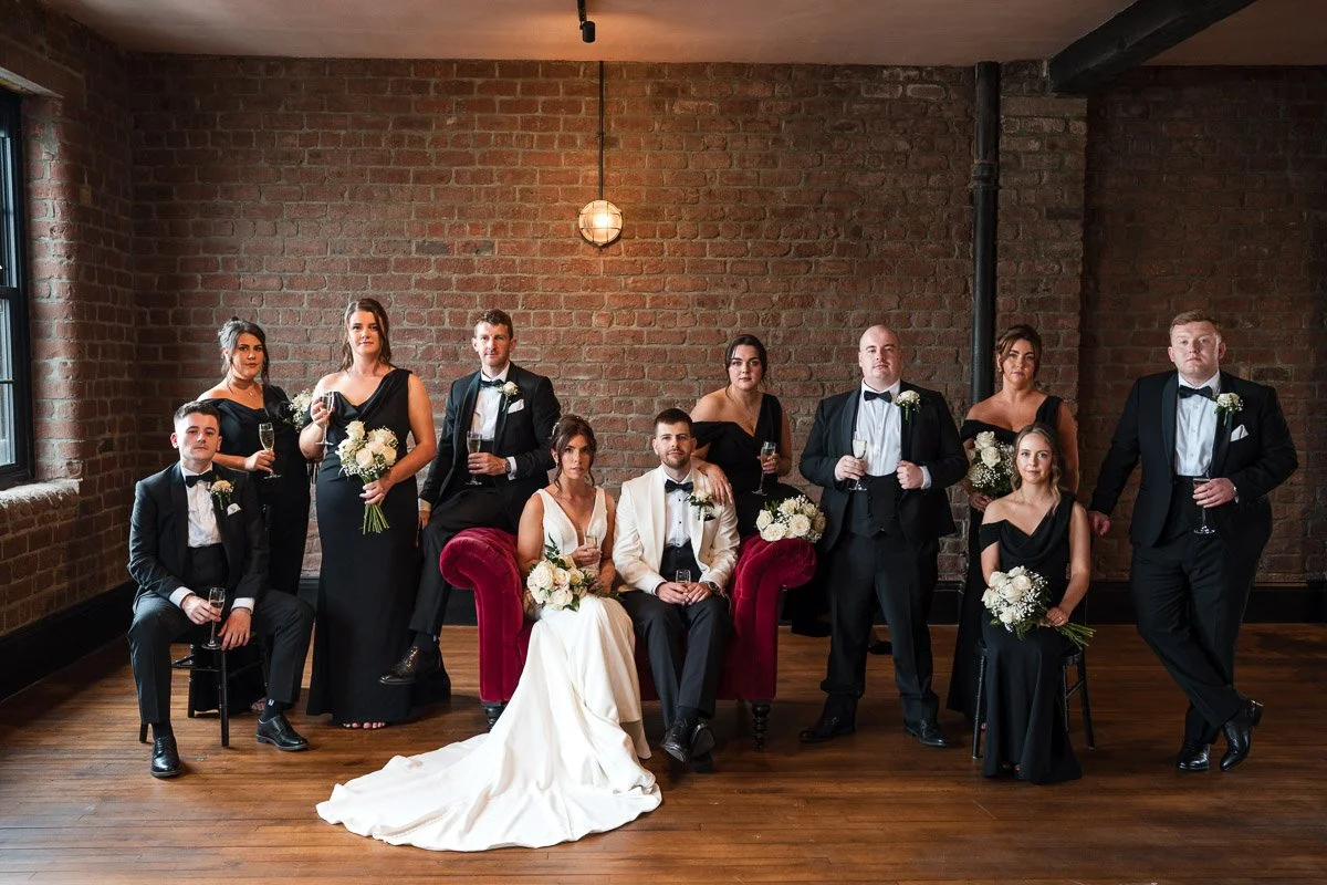 A wedding party with men in tuxedos and women in black dresses, some holding bouquets, posing in a brick-walled room with wooden floors and a red velvet sofa at Engine Works Glasgow