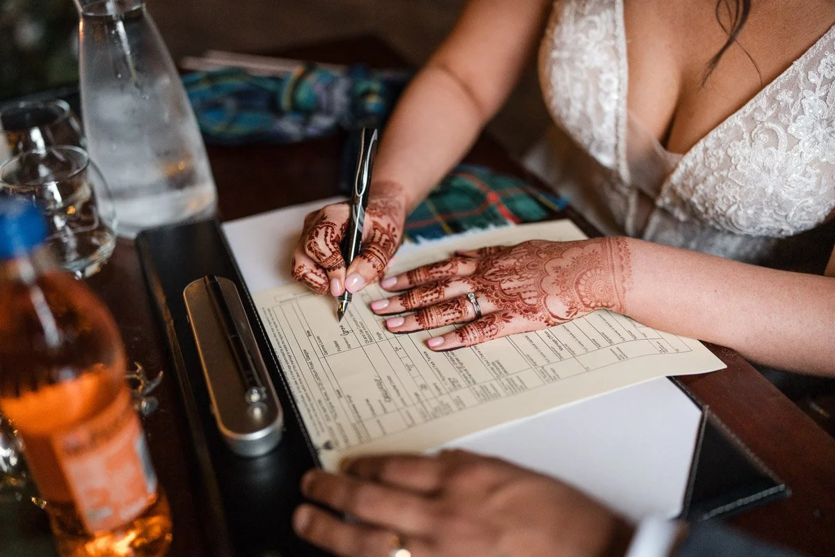 A woman with intricate henna tattoos on her hands and a wedding ring is signing a document at a wooden table, with water bottles and a small recorder nearby at Engine Works