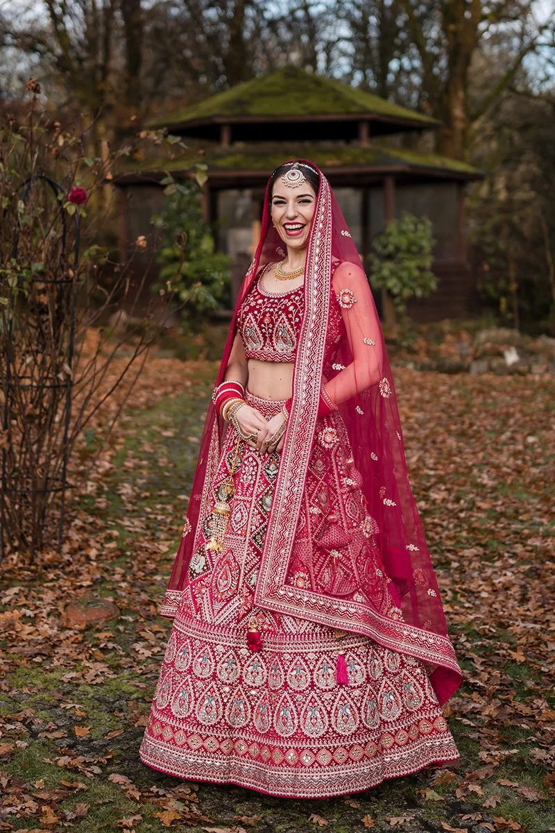A woman dressed in a traditional red Indian wedding attire stands outdoors among autumn leaves, smiling, with a wooden structure and trees in the background before her Hindu wedding in Glasgow
