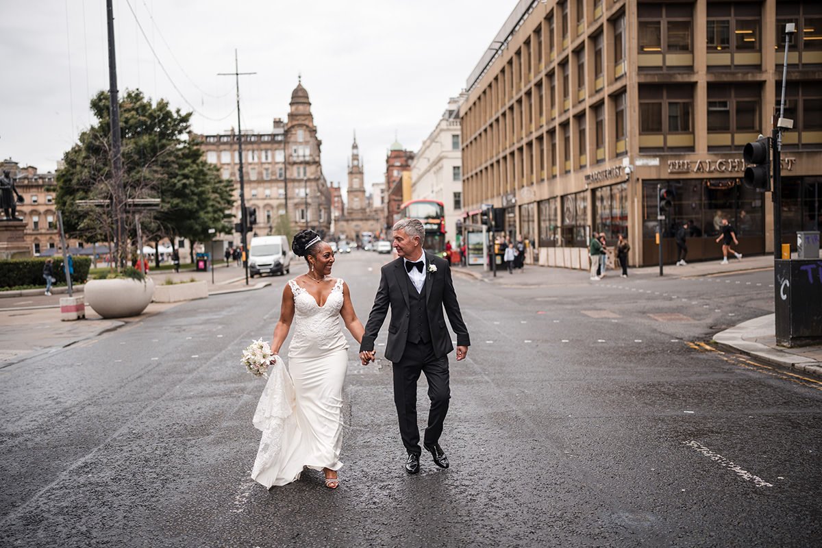 A newlywed couple holding hands and walking across a Glasgow street, with the bride in a white wedding dress holding a bouquet and the groom in a black tuxedo, after a city centre elopement in Glasgow