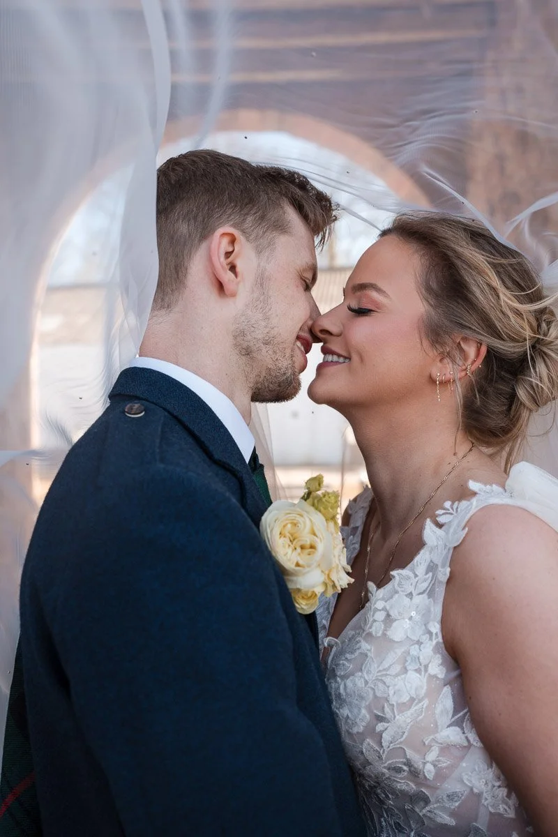 A bride and groom about to kiss, inside a transparent wedding veil, with the bride holding a bouquet of white roses at Enterkine House Hotel