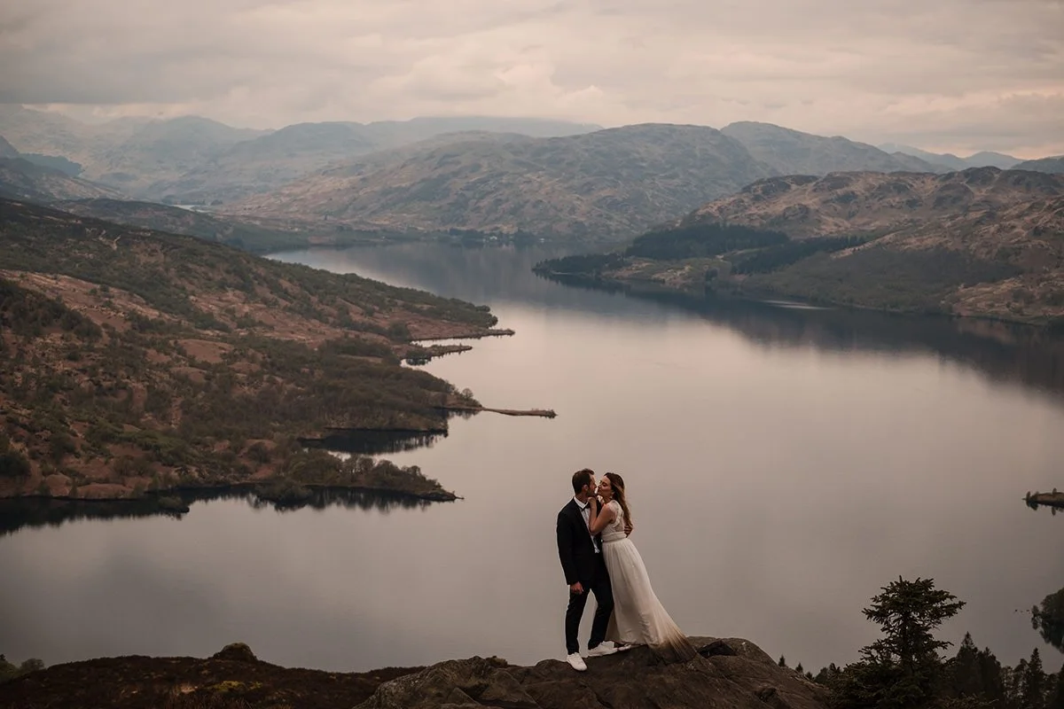 A bride and groom dressed in wedding attire standing on a rock overlooking a large loch with mountains in the background after their elopement on Ben A'an overlooking Loch Katrine