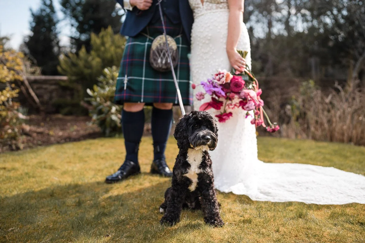 A black and white dog sitting on grass in front of a wedding couple, with a woman holding a bouquet of flowers, and a man dressed in traditional Scottish attire in the background at Loch Ness House Hotel