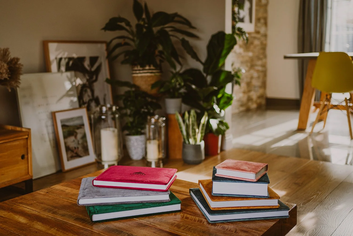 Stacked notebooks with velvet covers on a wooden table in a cozy living room with plants and artwork in the background.
