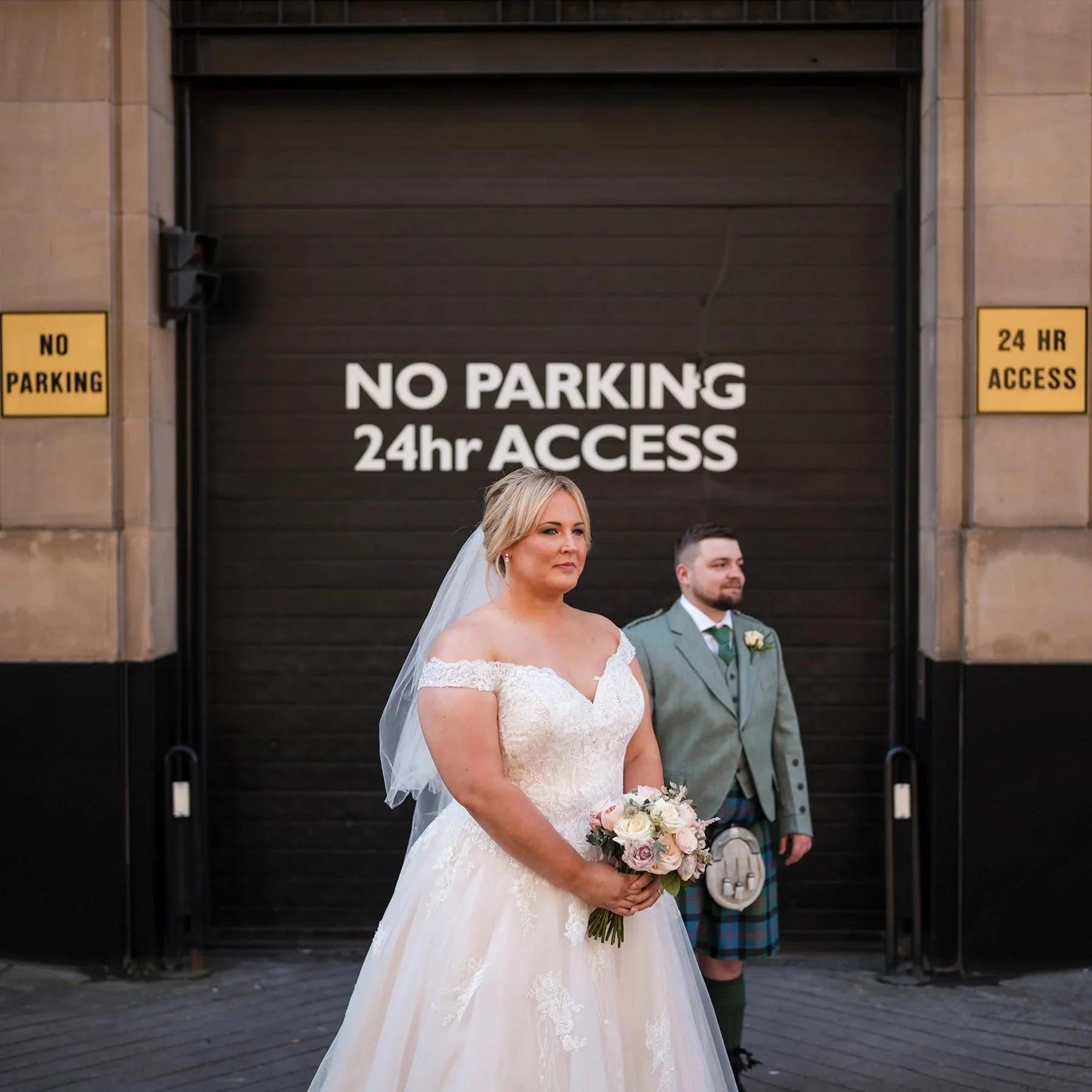 A bride in a wedding dress holding a bouquet standing in front of a black door with a sign that reads "NO PARKING 24hr ACCESS." A groom in a kilt and suit stands behind her, slightly to the right, in front of the door.