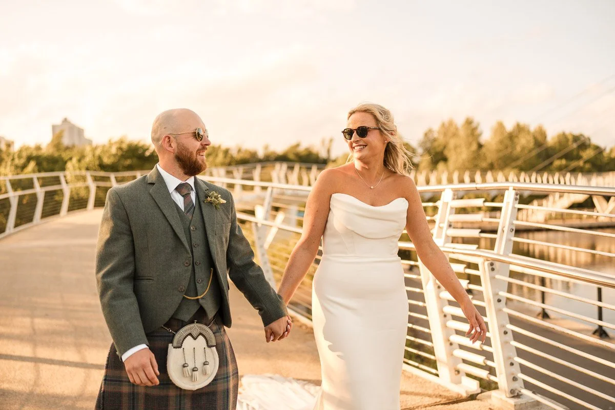 A couple dressed in wedding attire holding hands and smiling on a modern footbridge during sunset, with trees in the background at their Engine Works wedding