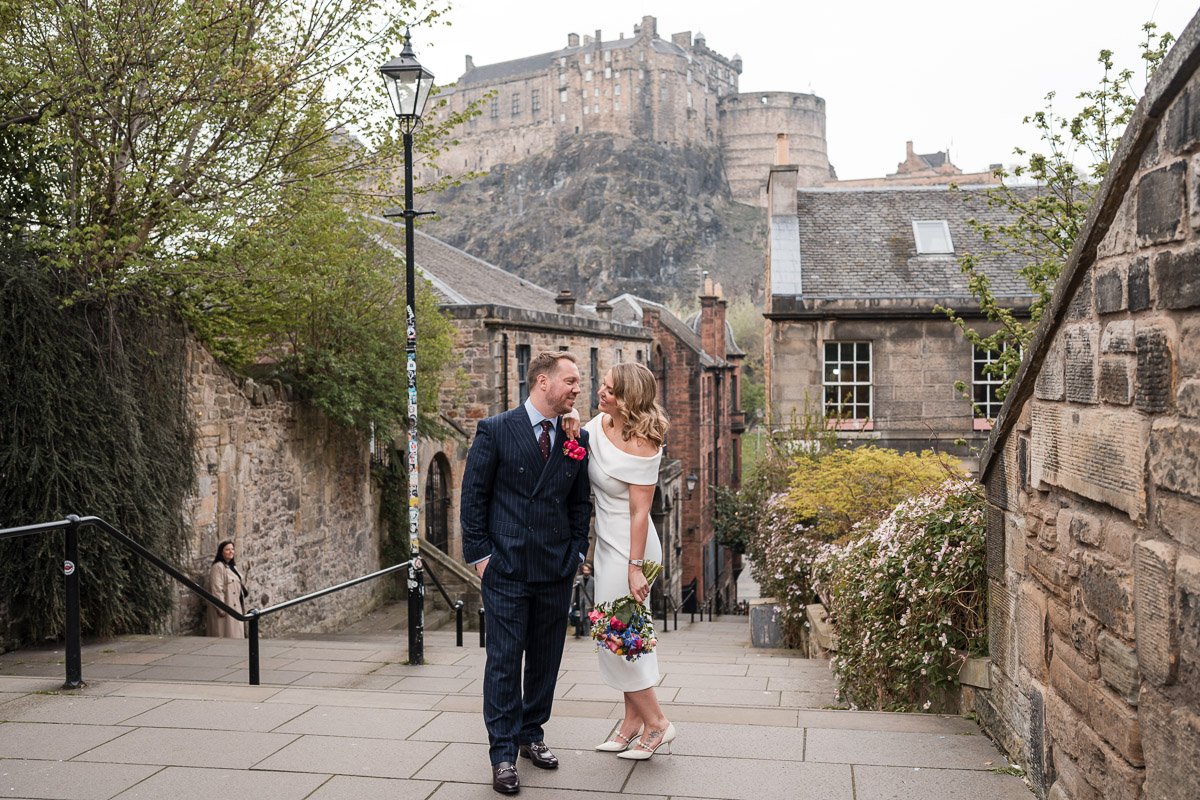 A couple in wedding attire standing on a city street with Edinburgh Castle in the background. The man is in a navy pinstripe suit and the woman is in a white dress holding flowers. They are looking at each other smiling.