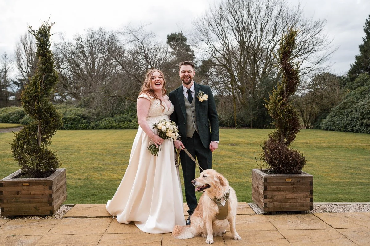 Bride and groom standing outdoors on a cloudy day, smiling with a golden retriever sitting in front of them. The bride is holding a bouquet of white flowers and wearing a long white wedding dress, while the groom is in a dark suit with a boutonniere.