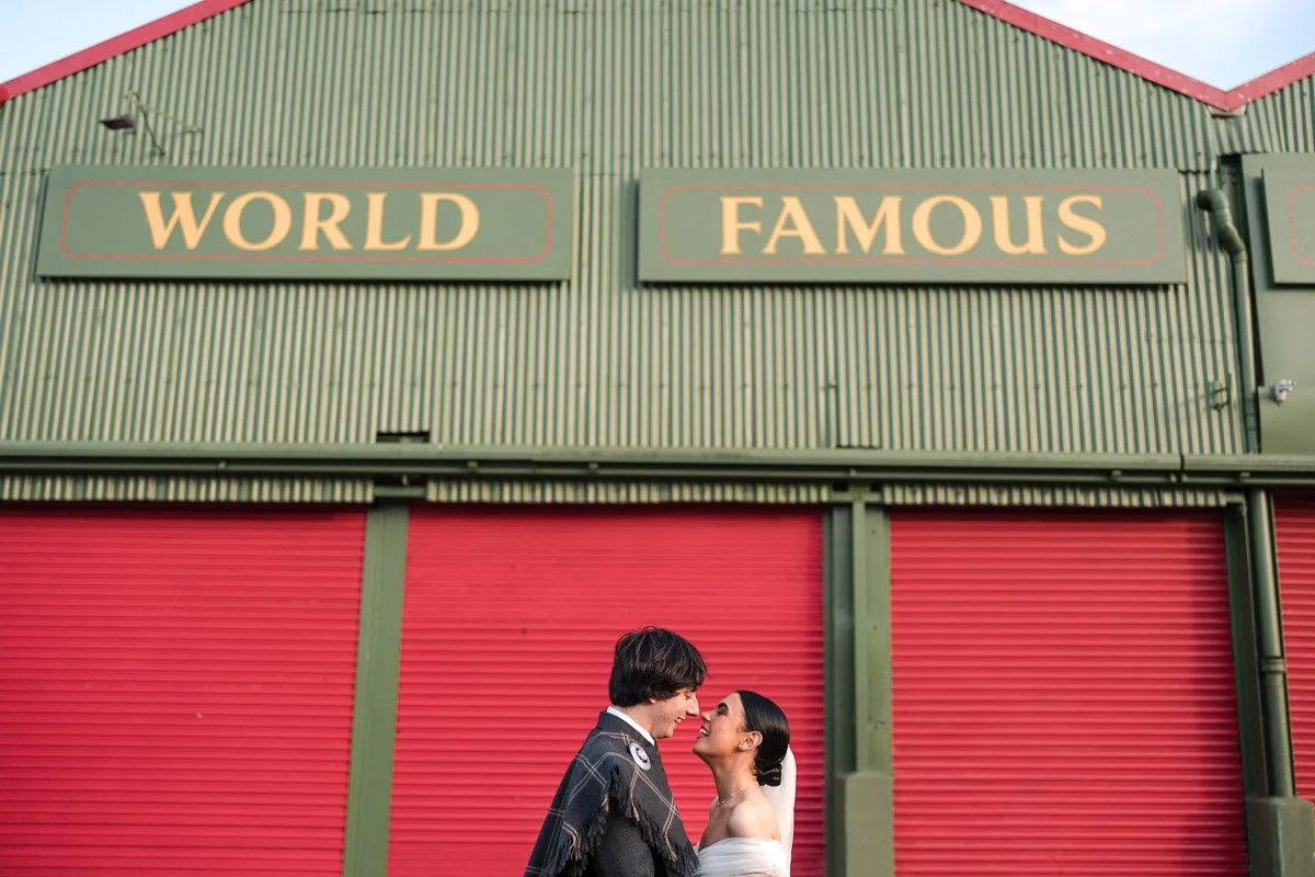 A bride and groom in wedding attire standing close and smiling in front of a green building with red doors and signs that say "WORLD FAMOUS" at Barras Market, Glasgow.