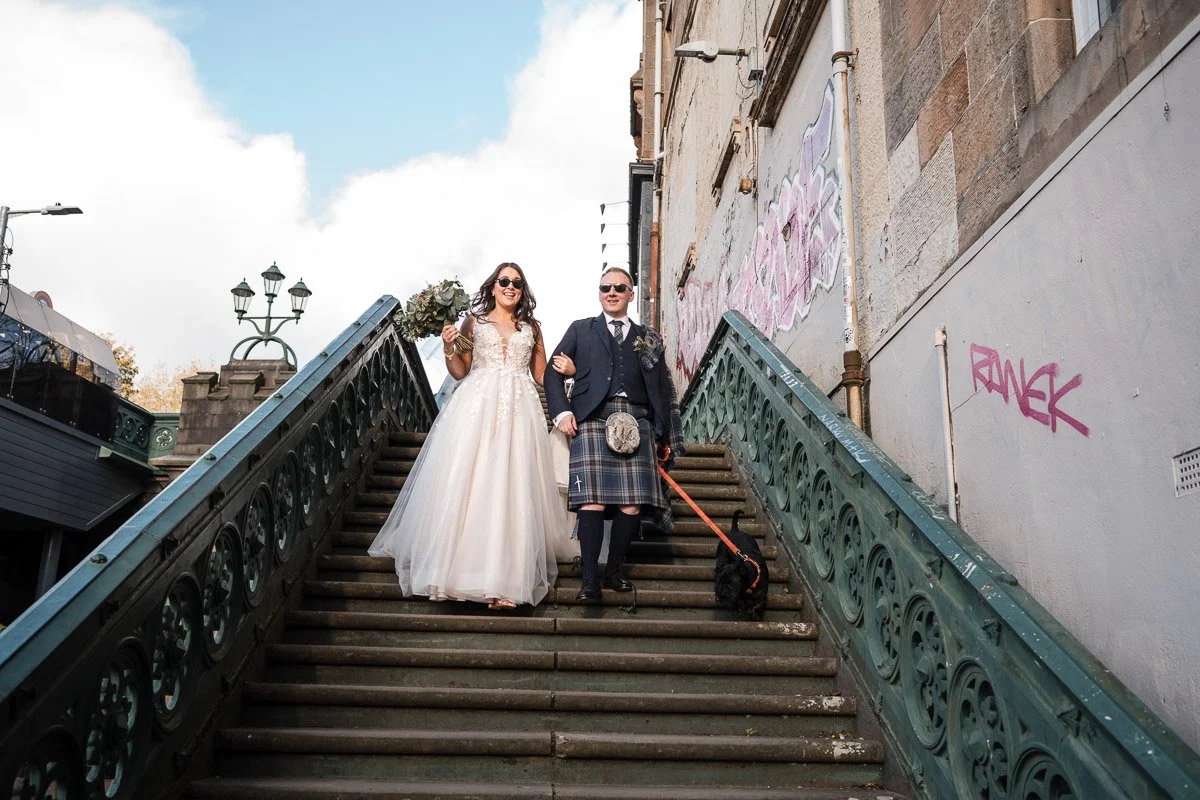A bride and groom walking down outdoor stairs, holding hands, with the bride carrying a bouquet, and the groom dressed in a tuxedo with a Scottish kilt, accompanied by a small dog on a leash in Glasgow West End