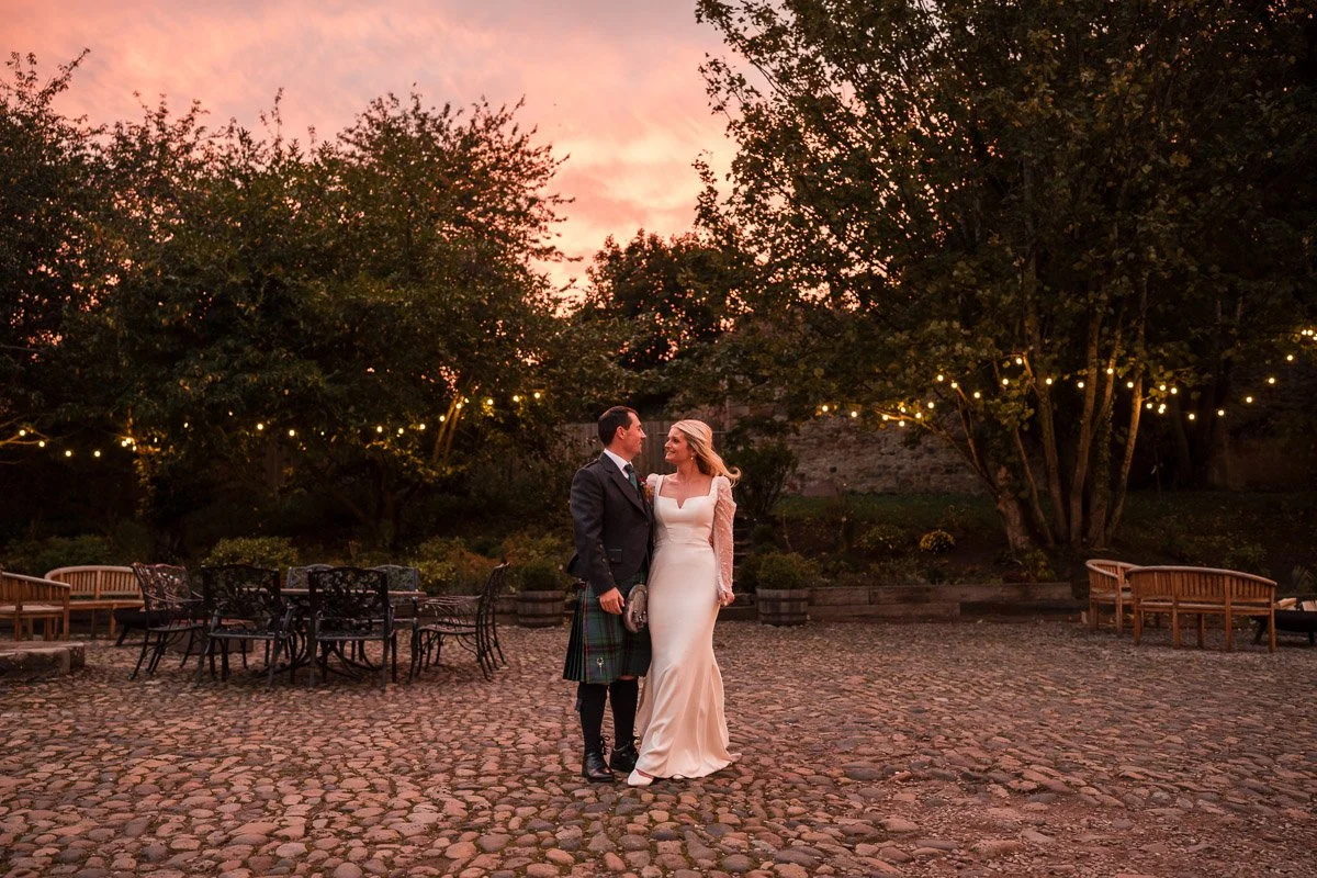 Bride and groom dancing outdoors during sunset with string lights, cobblestone ground, surrounded by trees and outdoor furniture at Broxmouth Courtyard