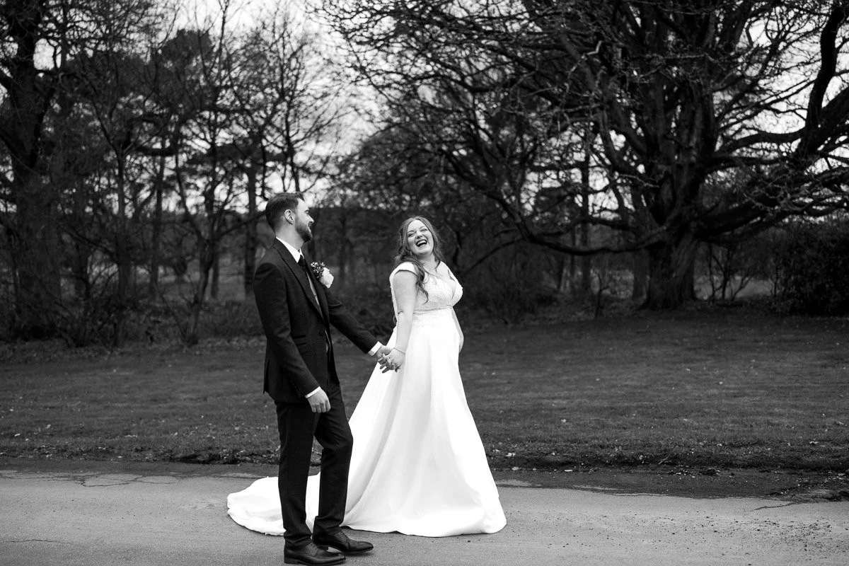 A black and white photo of a bride and groom holding hands outdoors, smiling and laughing. The bride wears a long white dress, and the groom wears a dark suit with a boutonniere. Behind them are leafless trees.