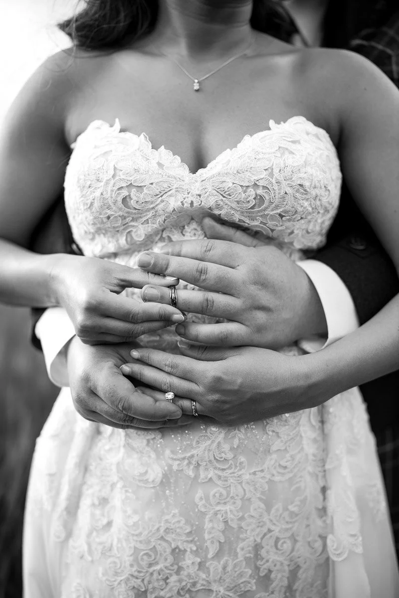 A close-up black and white photo of a bride in a lace wedding dress being embraced by a groom, with their hands and rings visible.