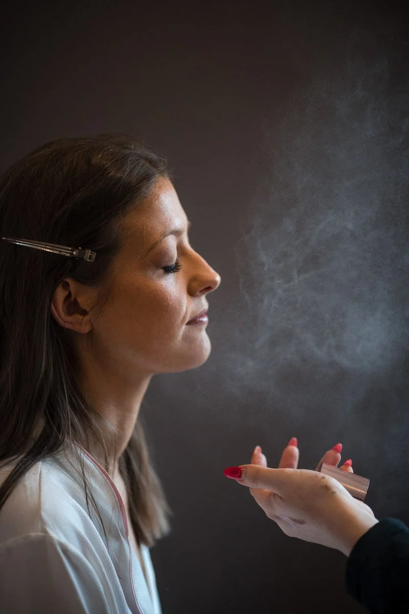 A bride with closed eyes is getting her face misted or sprayed with a water spray by a makeup artist with red nail polish at Brig o' Doon House Hotel