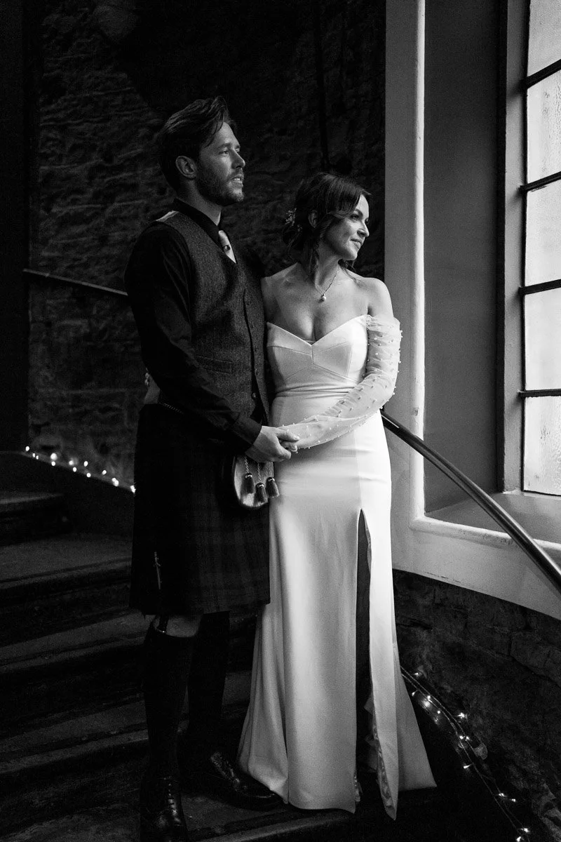 Black and white photo of a man and woman in wedding attire standing by a window, holding hands, with the woman wearing a strapless gown and the man in traditional Scottish kilt and vest, looking out the window.