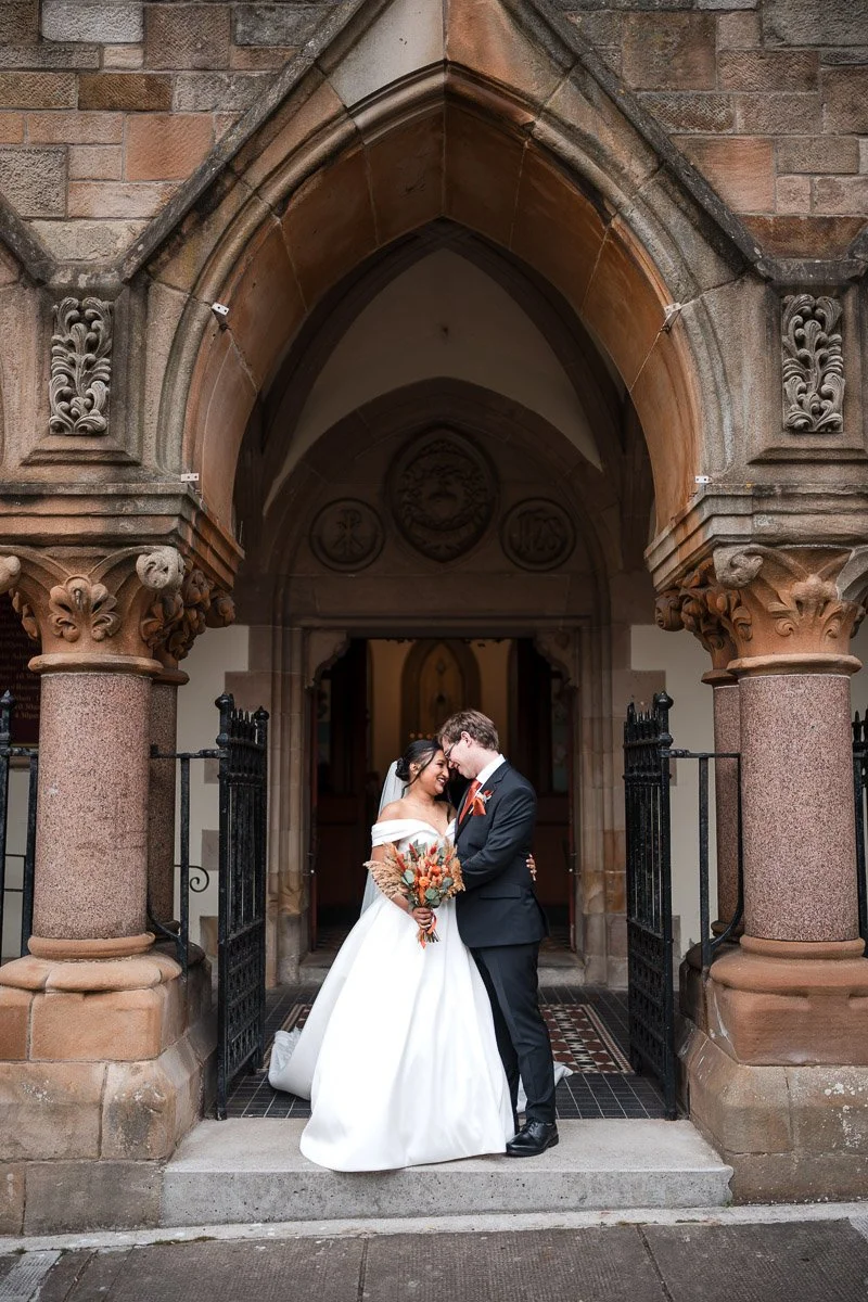 A wedding couple standing in front of a church entrance, embracing and smiling, with a bride in a white gown holding a bouquet, and a groom in a black suit with an orange tie at a Glasgow wedding