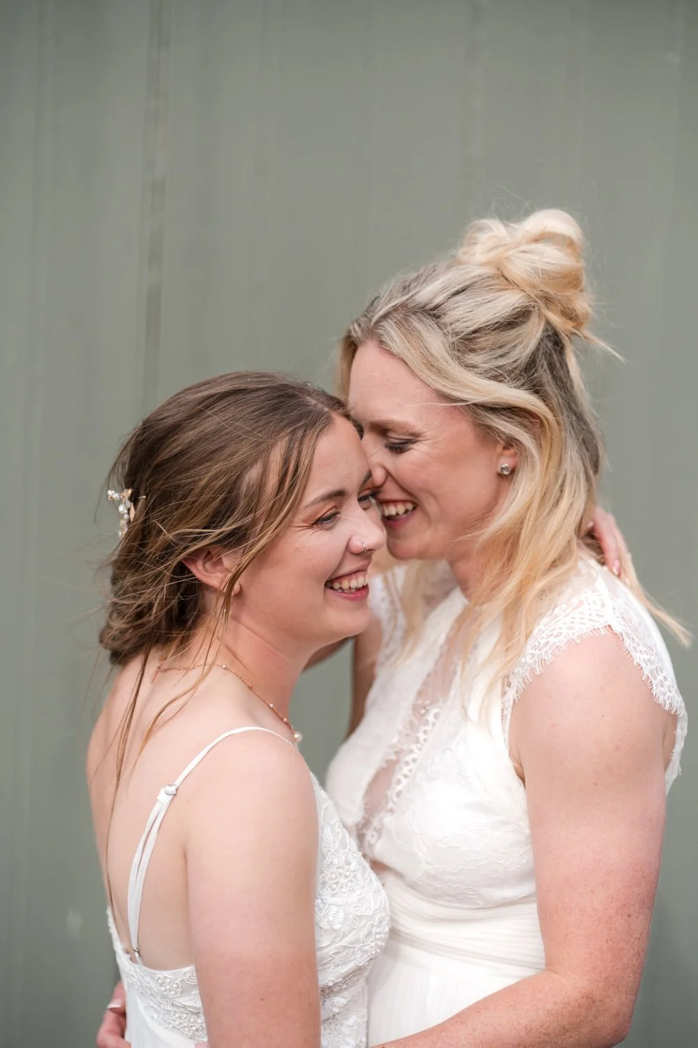 Two brides, dressed in white, are smiling and embracing each other closely, forehead to forehead, in an affectionate moment at Cow Shed Crail