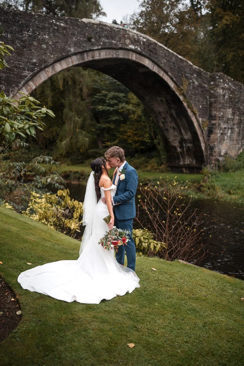 A bride and groom sharing a kiss outdoors with a stone bridge in the background and lush greenery surrounding them at Brig o' Doon