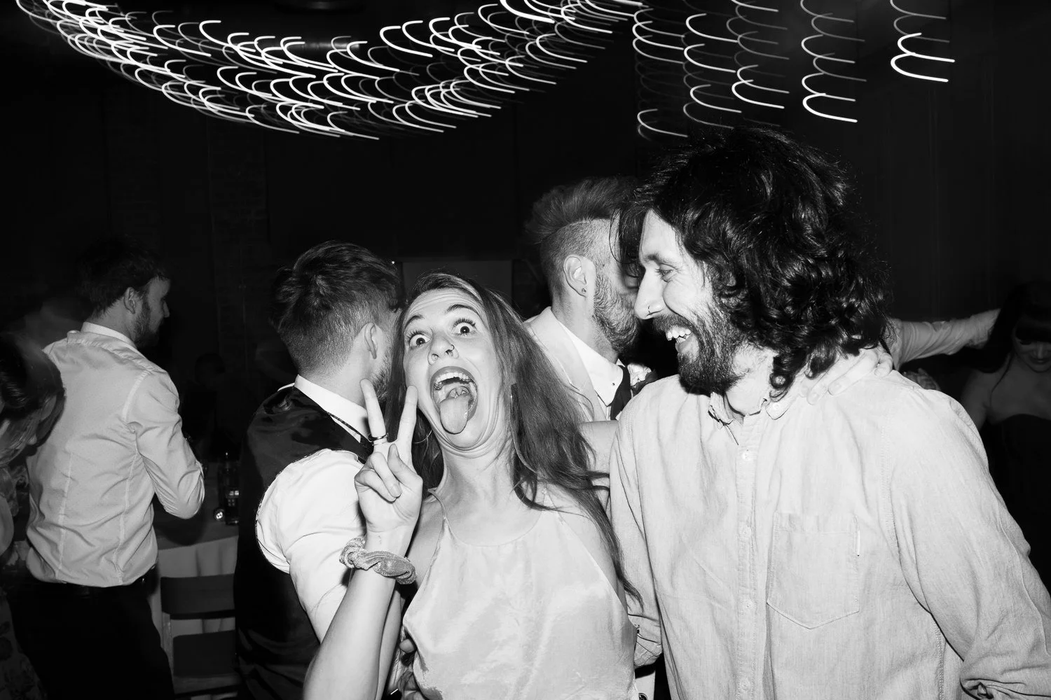 A black-and-white photo of a group of people at a party, with two people in the foreground laughing and posing for the camera. The woman is making a peace sign with her fingers and sticking out her tongue, and the man is smiling widely.