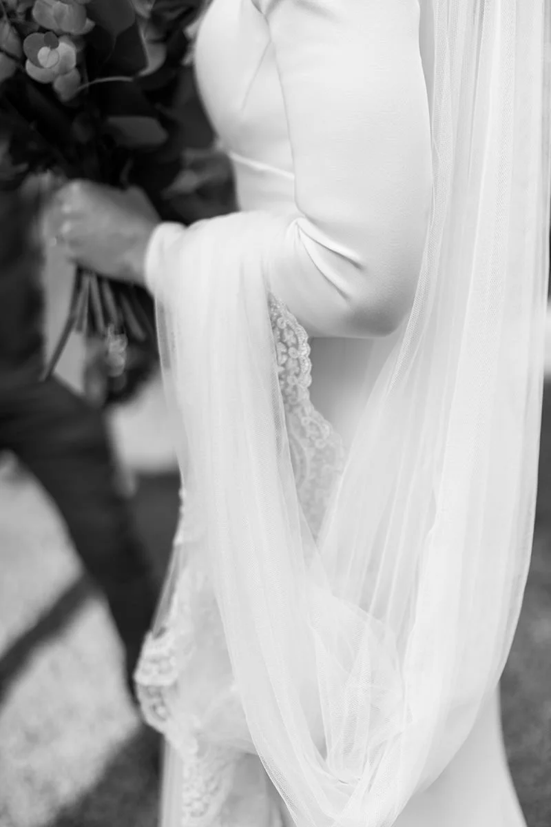 Close-up of a Sassi Holford wedding dress showing the bride’s veil draped over the lace sleeve while she holds her bouquet