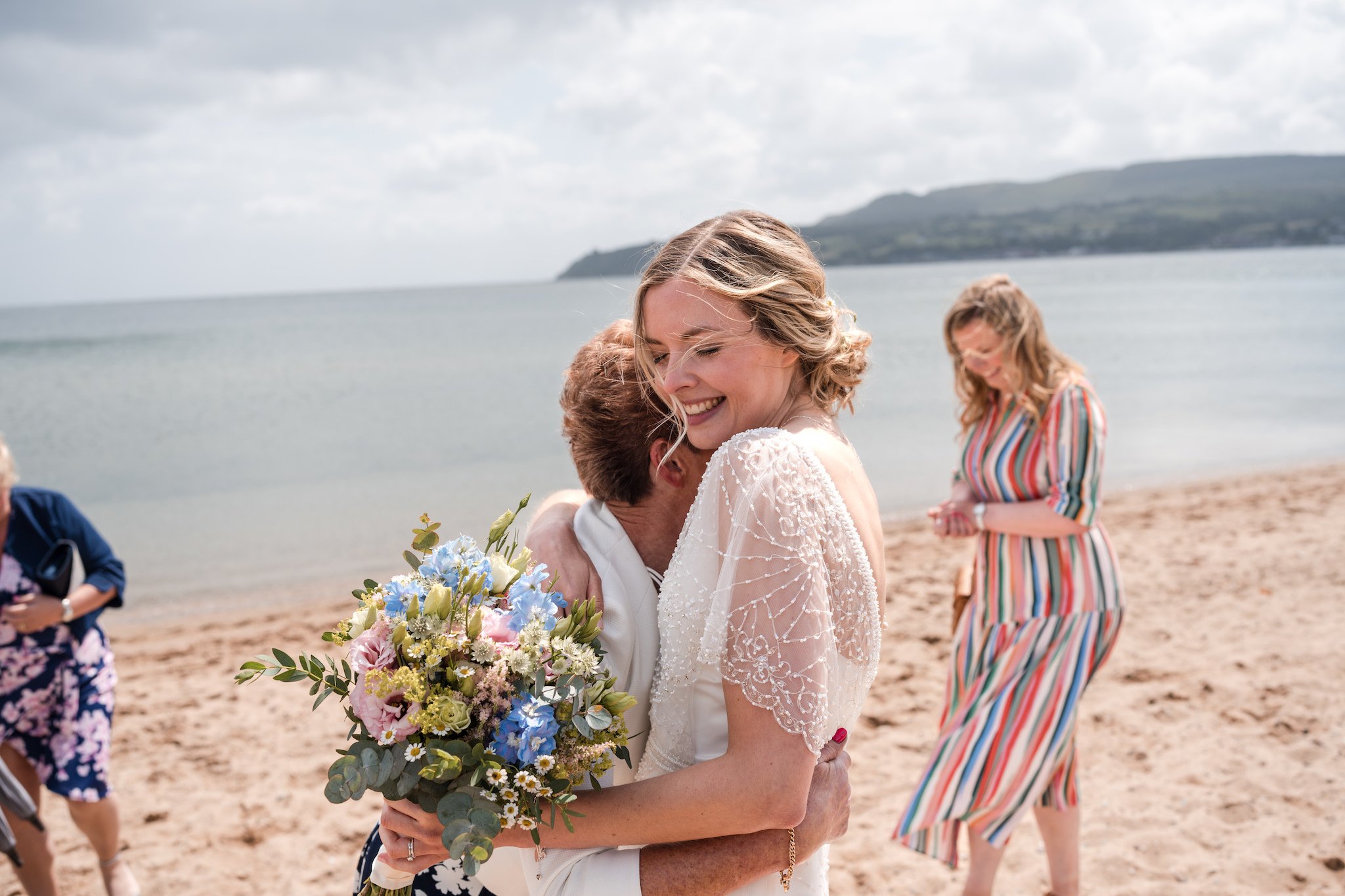 A bride hugging a guest on the beach, holding a bouquet of flowers, with other guests in the background at an elopement in Arran