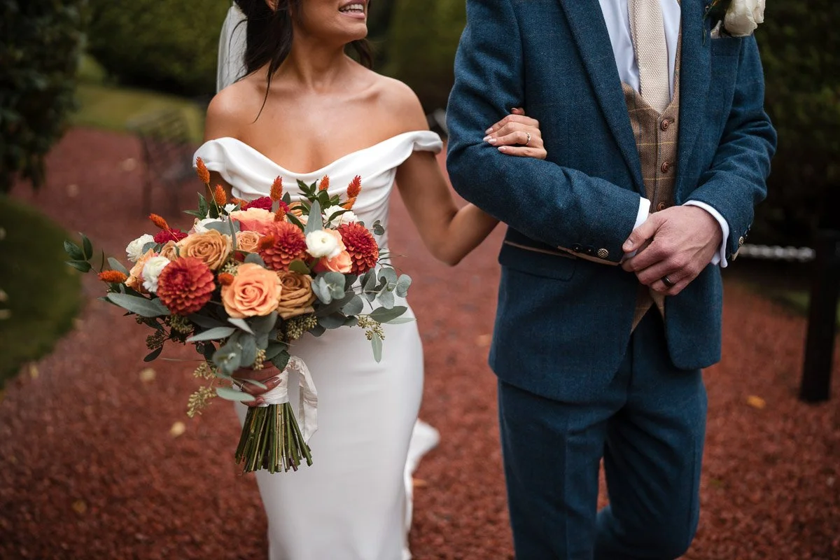 Bride holding a bouquet of flowers while walking arm-in-arm with her groom, during an outdoor wedding at Brig o' Doon