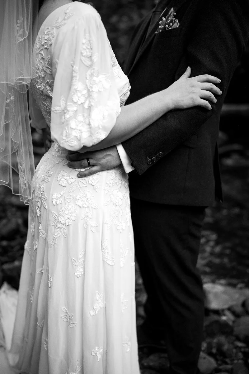 A black and white photo of a bride and groom holding each other at their wedding, with the bride in a detailed lace wedding dress and the groom in a dark suit.