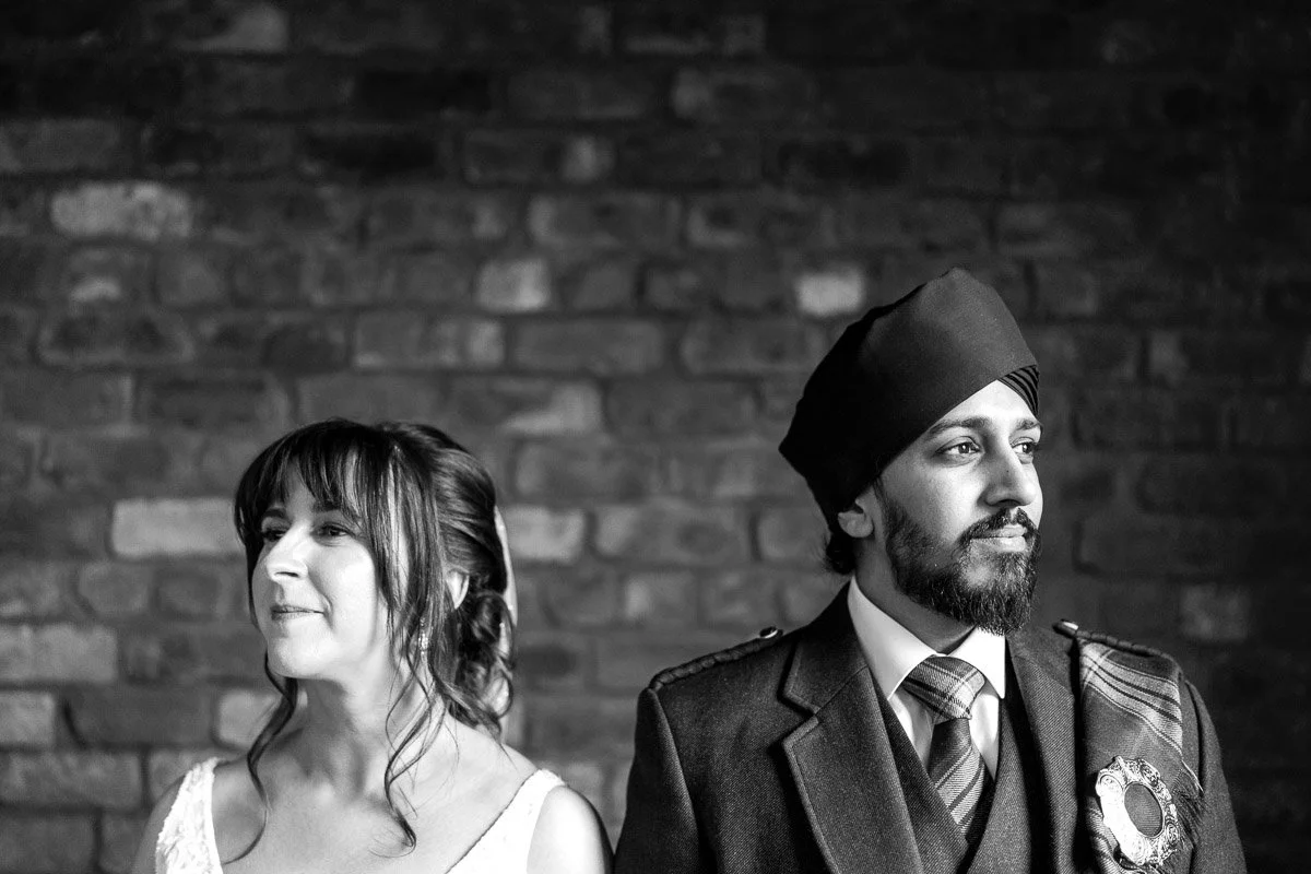 Woman with shoulder-length hair wearing a white dress and a man with a beard wearing a turban and kilt, standing against a brick wall at Engine Works, Glasgow