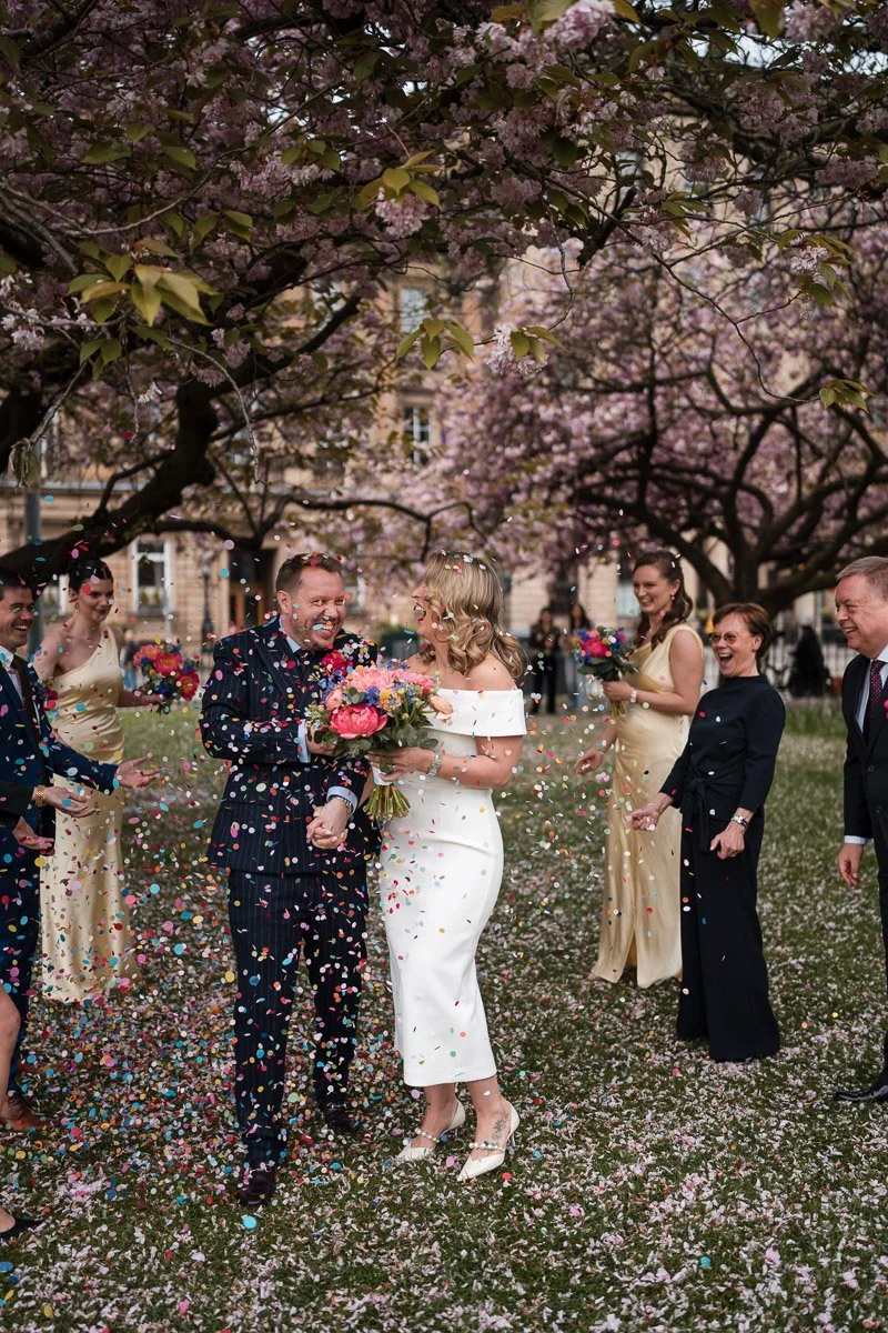 People celebrating a wedding under blooming pink cherry blossom trees, surrounded by colourful confetti and holding bouquets after a wedding at Gleneagles Town House Edinburgh