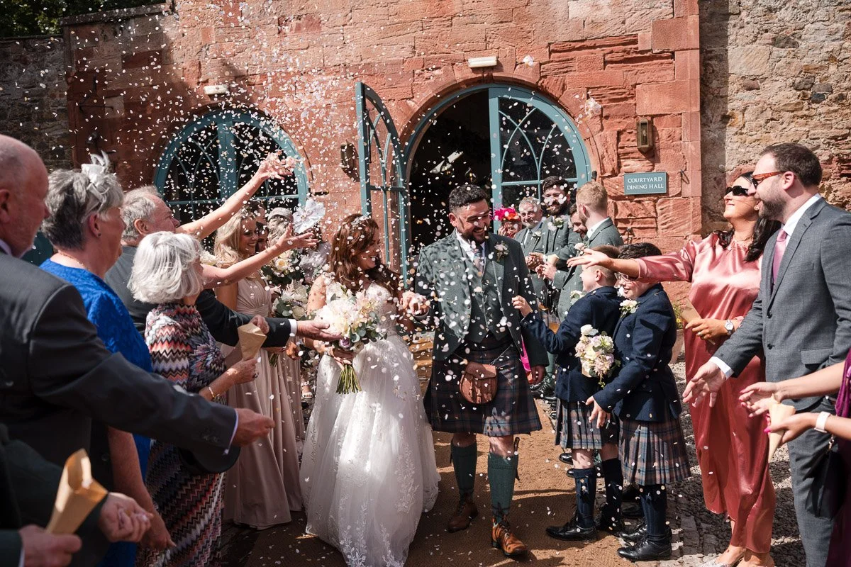 A wedding celebration with a bride and groom walking through a crowd of guests outside a brick building. The guests are throwing confetti and smiling at Broxmouth Courtyard
