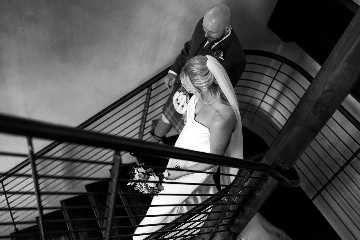 Black and white photo of a bride and groom descending a spiral staircase; the bride is wearing a strapless wedding gown with a veil and holding a bouquet of flowers, while the groom is dressed in a formal jacket and kilt at Engine Works