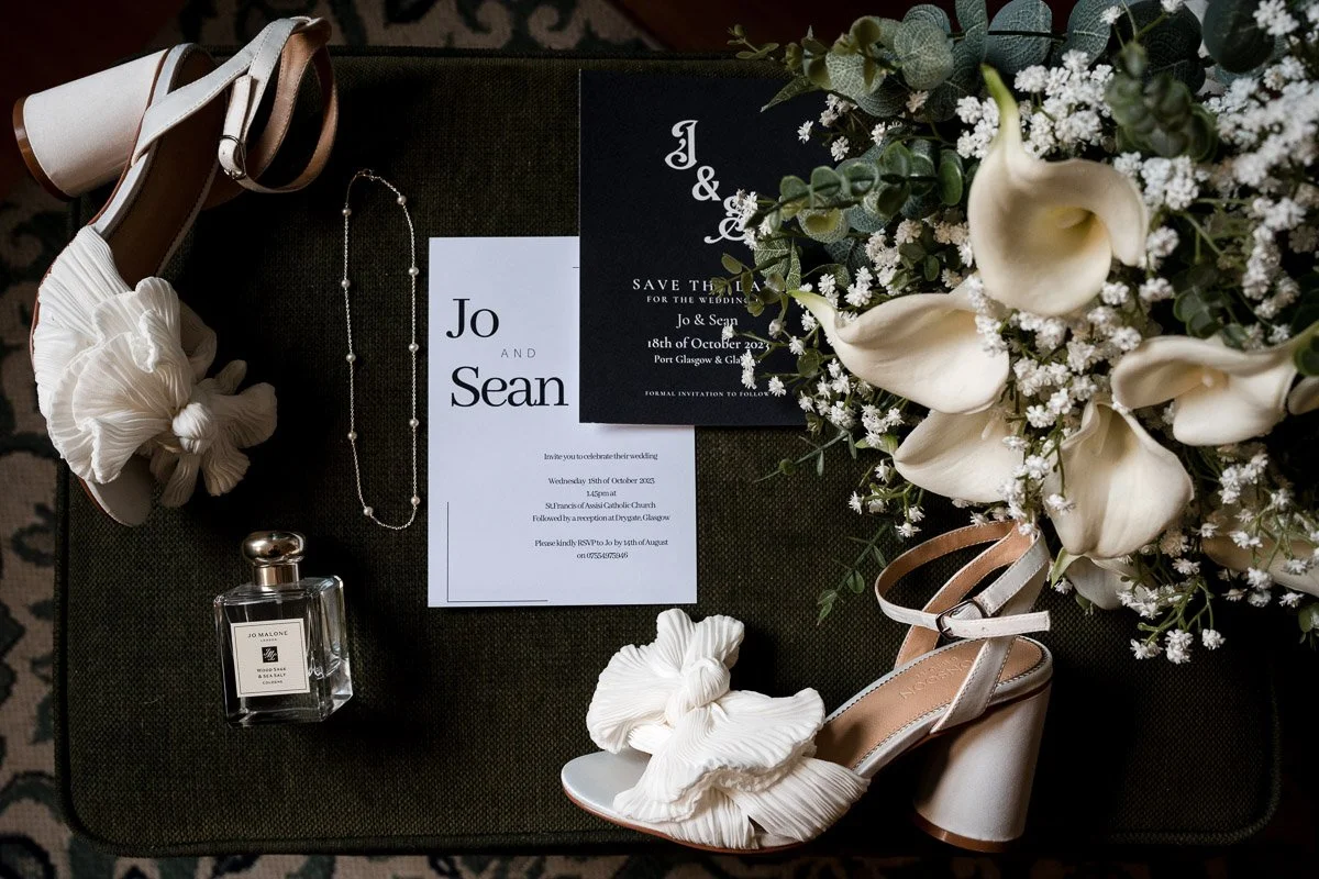 Wedding invitation, pearl necklace, perfume bottle, high-heeled shoes, flower bouquet with white calla lilies and greenery, save the date card, and a scrunchie on a dark surface.