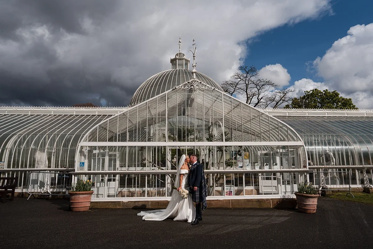 A bride and groom kissing in front of a large glass greenhouse at Botanical Gardens, Glasgow, with cloudy sky in the background.