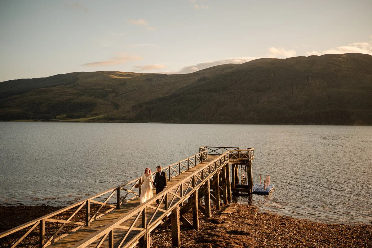 Wedding couple walking on a wooden dock by a lake during sunset with mountains in the background at Cruin Loch Lomond
