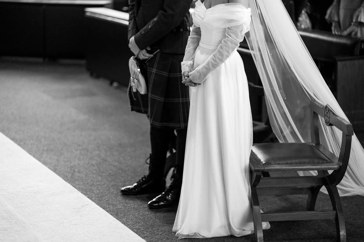 A couple dressed in wedding attire standing at the altar during a wedding ceremony. The bride is wearing a long white gown with sheer sleeves and a veil, while the groom is dressed in a dark suit and plaid kilt. There is a wooden chair and some drape