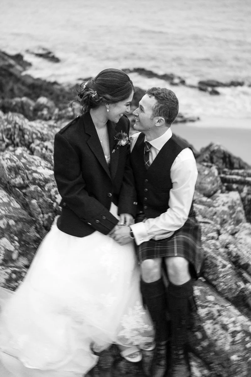 A black and white photo of a bride and groom, dressed in wedding attire, sitting on rocks by the ocean, embracing and smiling at each other at GG's Yard