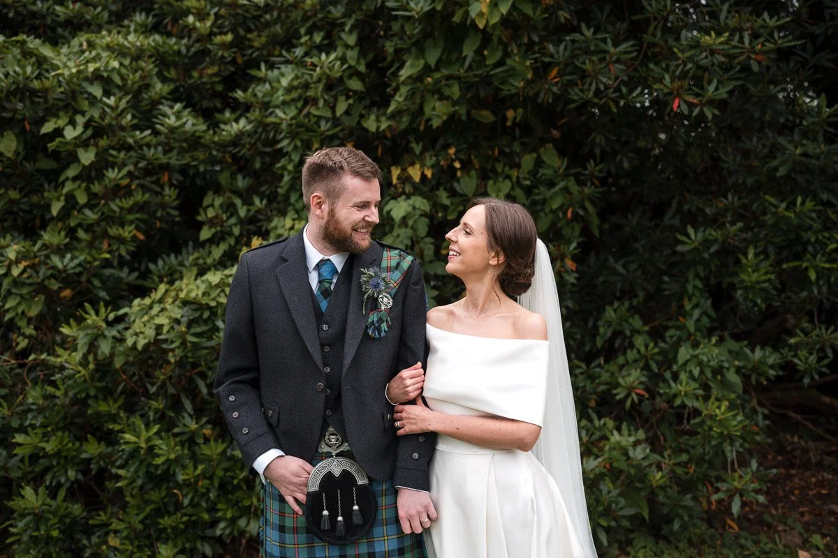 A bride and groom smiling and looking at each other outdoors with green bushes in the background. The groom wears a kilt and the bride wears a white off-the-shoulder wedding dress and veil at Boturich Castle