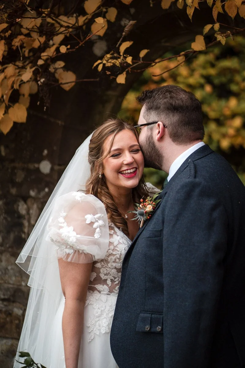 A bride and groom sharing a kiss outdoors during their wedding, with fall foliage in the background at Rufflets St Andrews