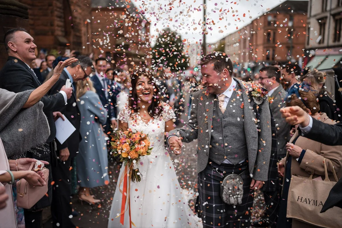 A bride and groom at their wedding celebration, smiling and holding hands as confetti falls around them, surrounded by friends and family in an outdoor urban setting in Glasgow