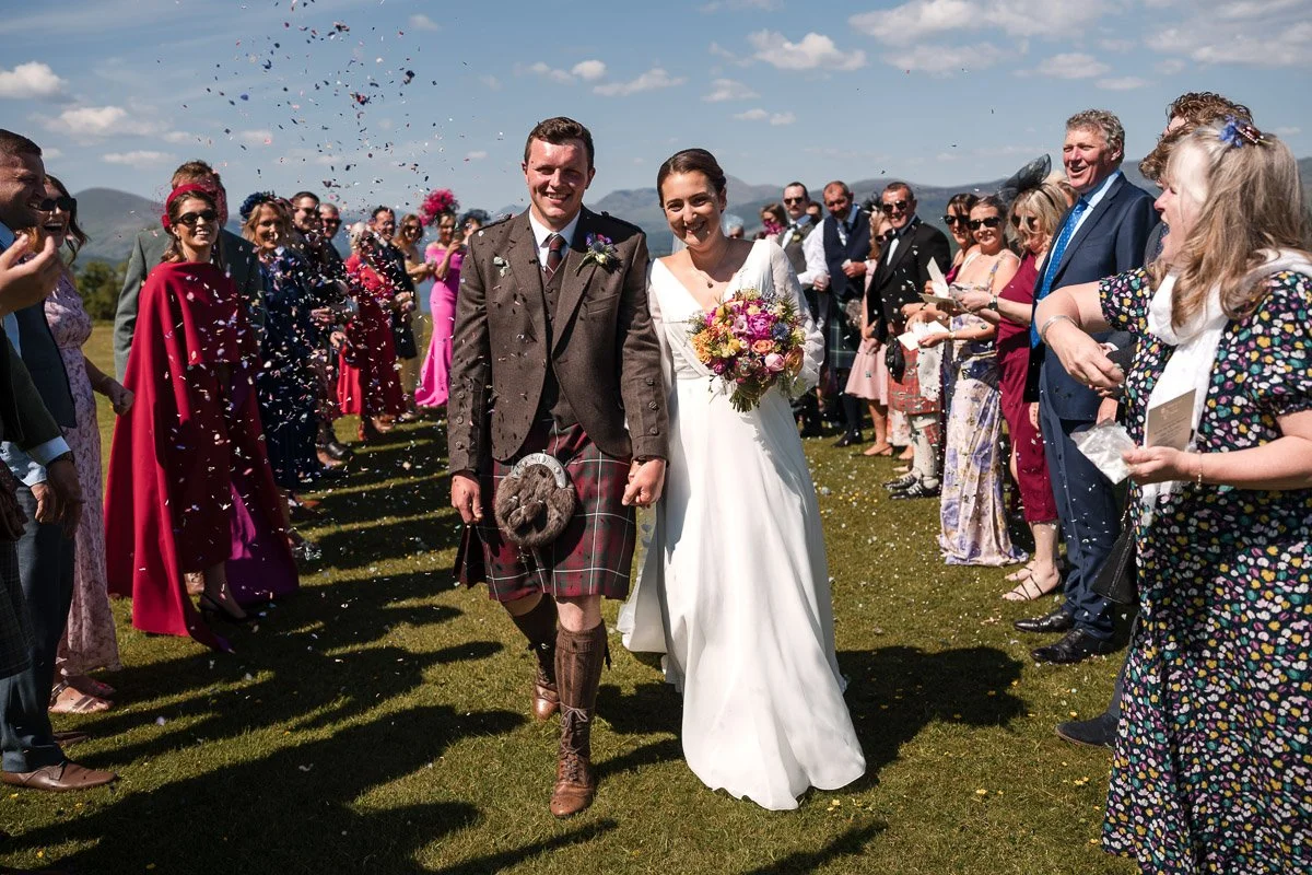 A bride and groom walking outdoors after wedding at Ardoch, Loch Lomond, surrounded by friends and family throwing confetti on a grassy field with mountains in the background.