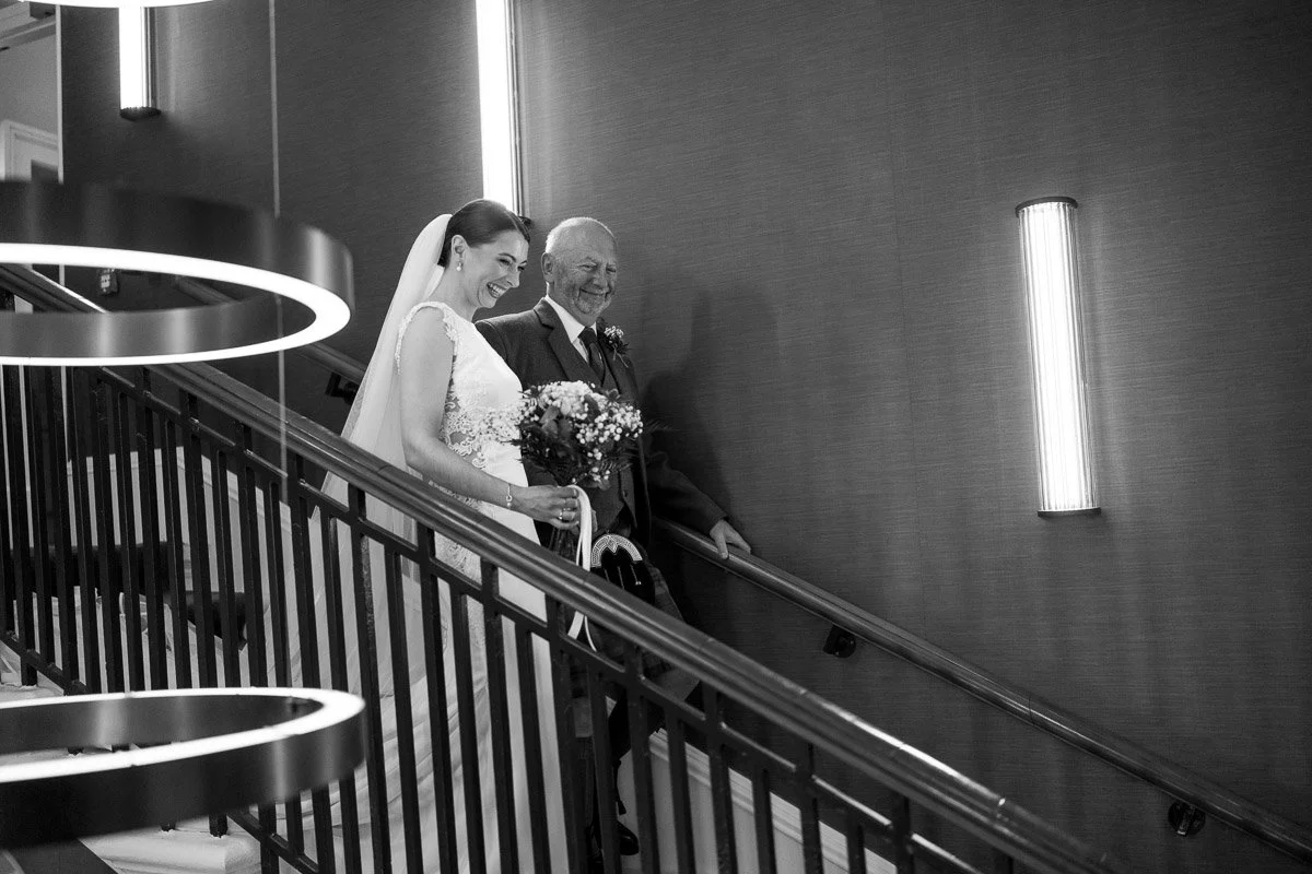 A bride and an her father, walking down a staircase at a wedding venue. The bride is smiling and holding a bouquet of flowers, wearing a wedding dress and veil, while the man is dressed in a suit before her Glasgow city elopement