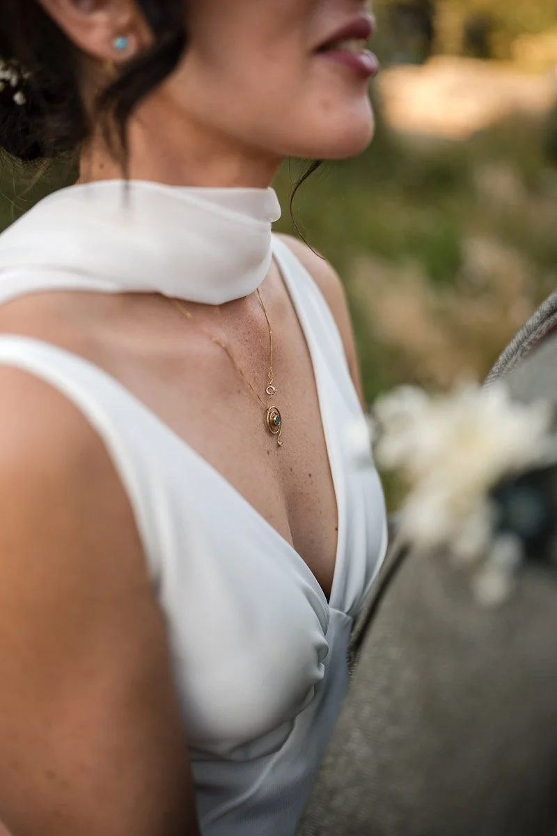 Close-up of a bride wearing a white sleeveless dress, a white scarf around her neck, a gold necklace with blue pendant, and turquoise earrings. The background is outdoors with blurred trees and sunlight at Kilmartin Castle