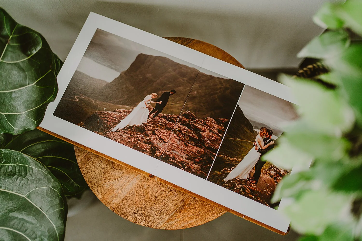 A photo album open to two wedding photos, featuring a bride and groom on a rocky landscape with green hills in the background, surrounded by green leaves.