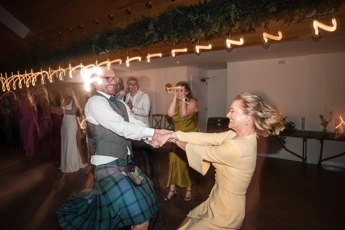A man wearing a kilt and a woman in a yellow dress dance and hold hands, smiling in a lively party setting with other guests in the background at a wedding at Broxmouth Courtyard