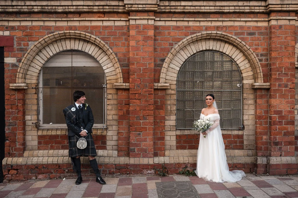 Bride in a white off-shoulder wedding gown holding a bouquet of white flowers, standing on the right side of a brick wall with metal grates over windows. Groom in traditional Scottish kilt and jacket, standing on the left side, looking at the bride.