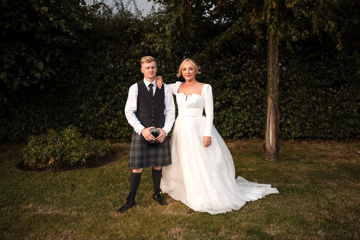 A woman in a white wedding dress and a man in traditional Scottish attire pose together outdoors on grass with trees in the background. The woman has blonde hair and is standing with her hand on the man's shoulder. The man is holding a camera and is 